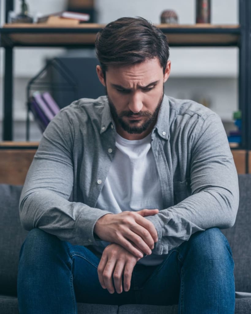 A man with a beard sits on a couch in Los Angeles, looking down with a thoughtful expression. He wears a gray shirt and jeans. Shelves with books on men's issues are in the background, hinting he might be a therapist for men.